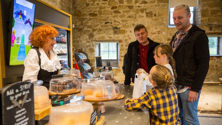 A woman with bright red hair is standing on one side of a café counter which is filled with cakes and bakes. To the right of the counter two male adults and